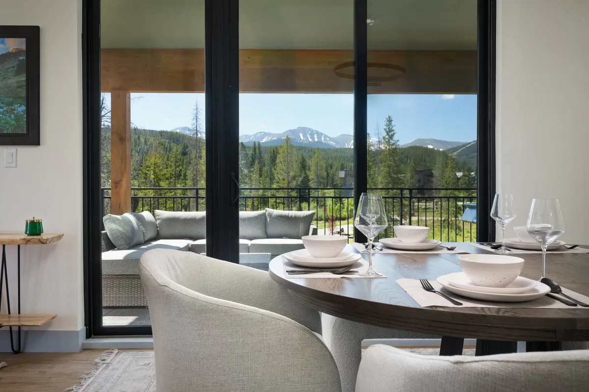 A beautifully set dining table with dishes and glassware positioned next to large windows that offer stunning mountain views. Comfortable outdoor seating is visible, blending indoor elegance with access to the outdoors.