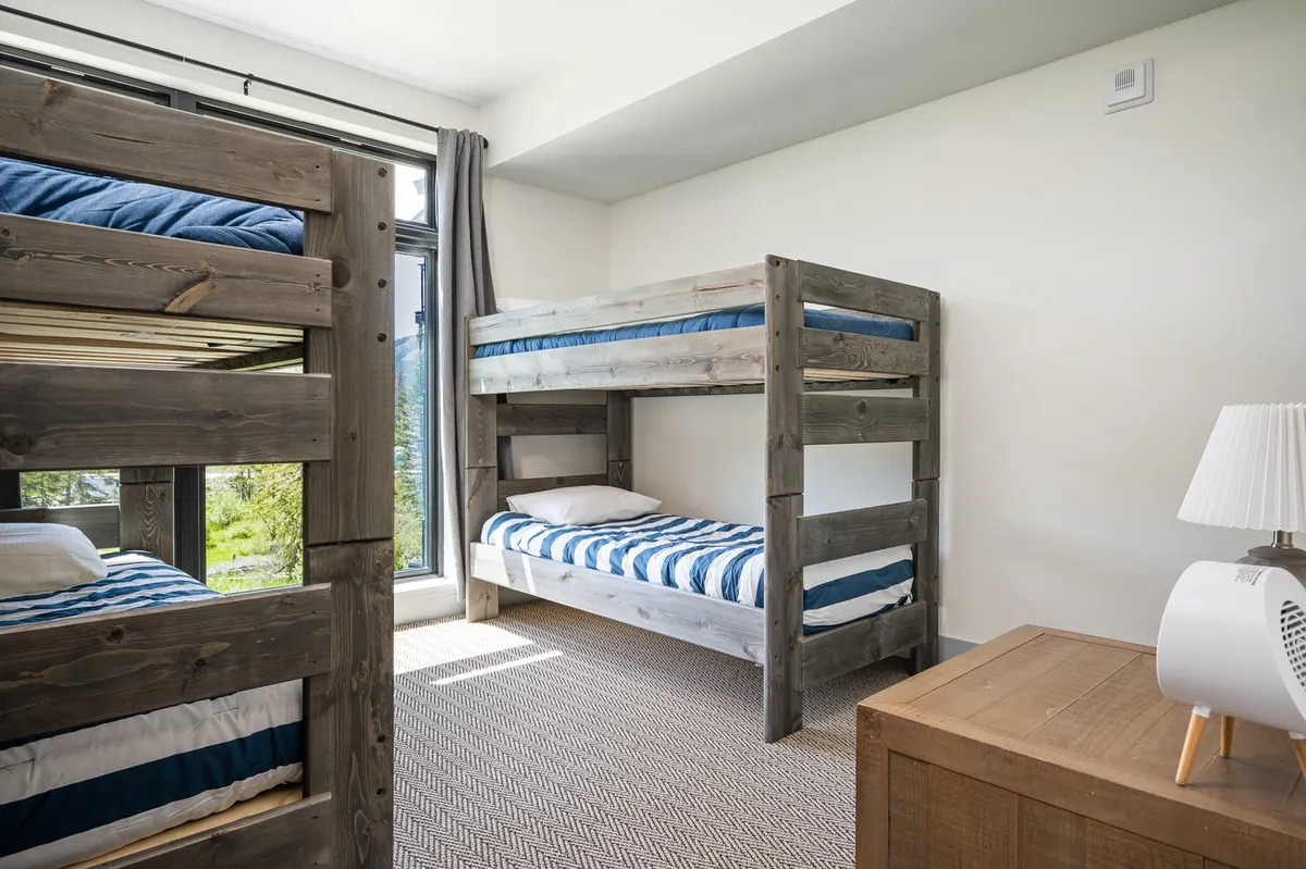 A cozy bunk room featuring two sets of twin-over-twin bunk beds, a window for natural light, and a nightstand. The playful yet inviting decor suggests a space designed for both children and groups, promoting fun and flexibility.