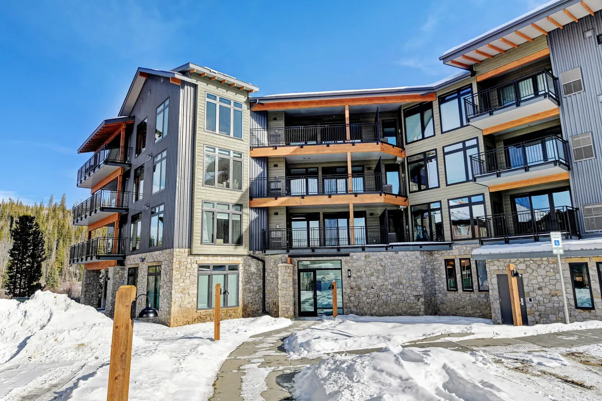 The image showcases the modern exterior architecture of a resort-style lodge, featuring large windows and balconies. The setting is welcoming and adventurous, hinting at nearby mountain activities and communal gatherings after a day of exploration.