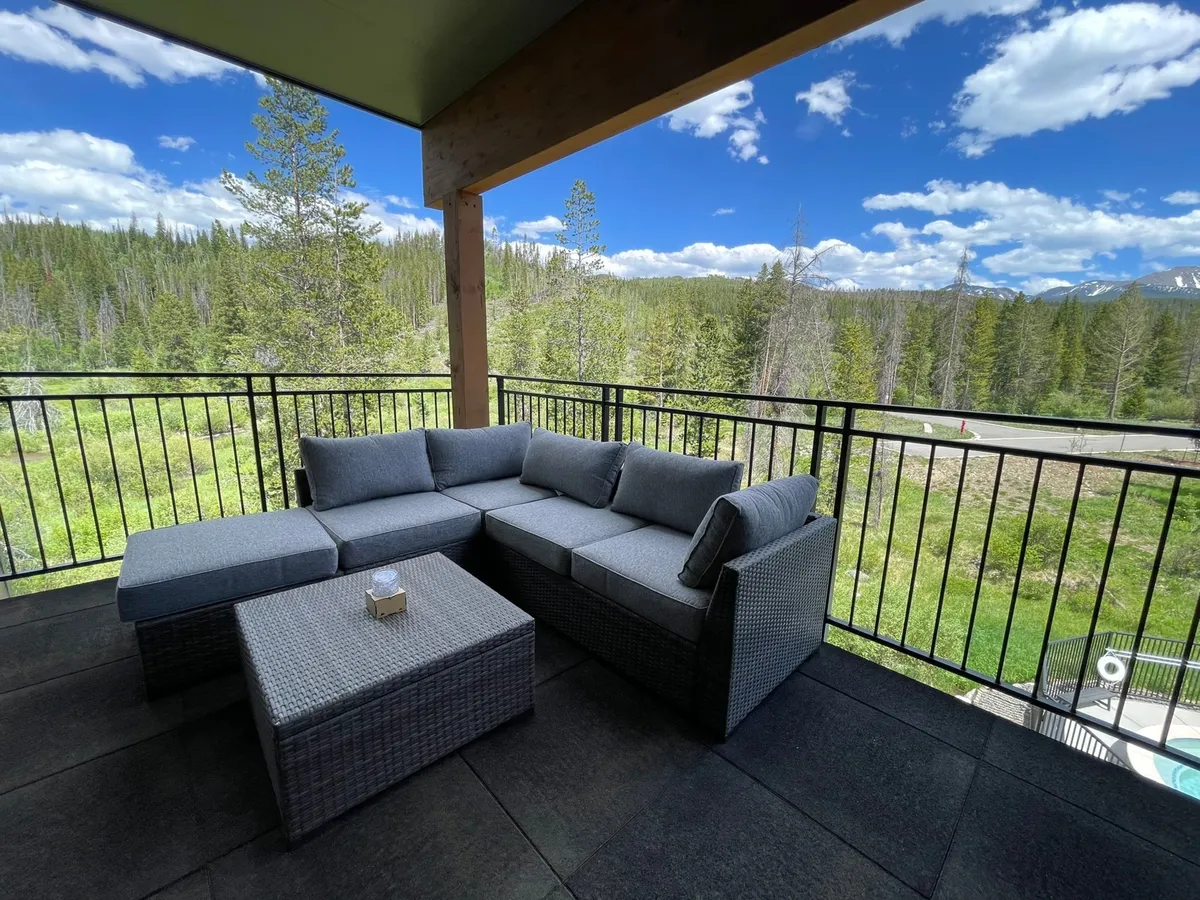 An inviting outdoor deck area featuring a sectional sofa and coffee table, surrounded by lush greenery and offering mountain views. The space is designed for relaxation, socializing, and enjoying the outdoors, with ample seating for family gatherings or quiet moments.