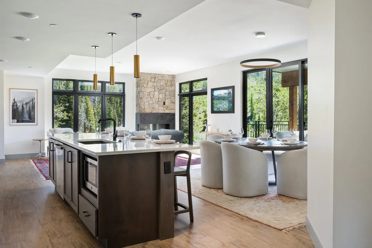 A beautifully appointed open-concept space featuring a sleek kitchen island and adjacent dining table, both set to host family meals and gatherings. The stone fireplace provides a cozy focal point, while large windows flood the room with natural light and offer views of the surrounding forest.