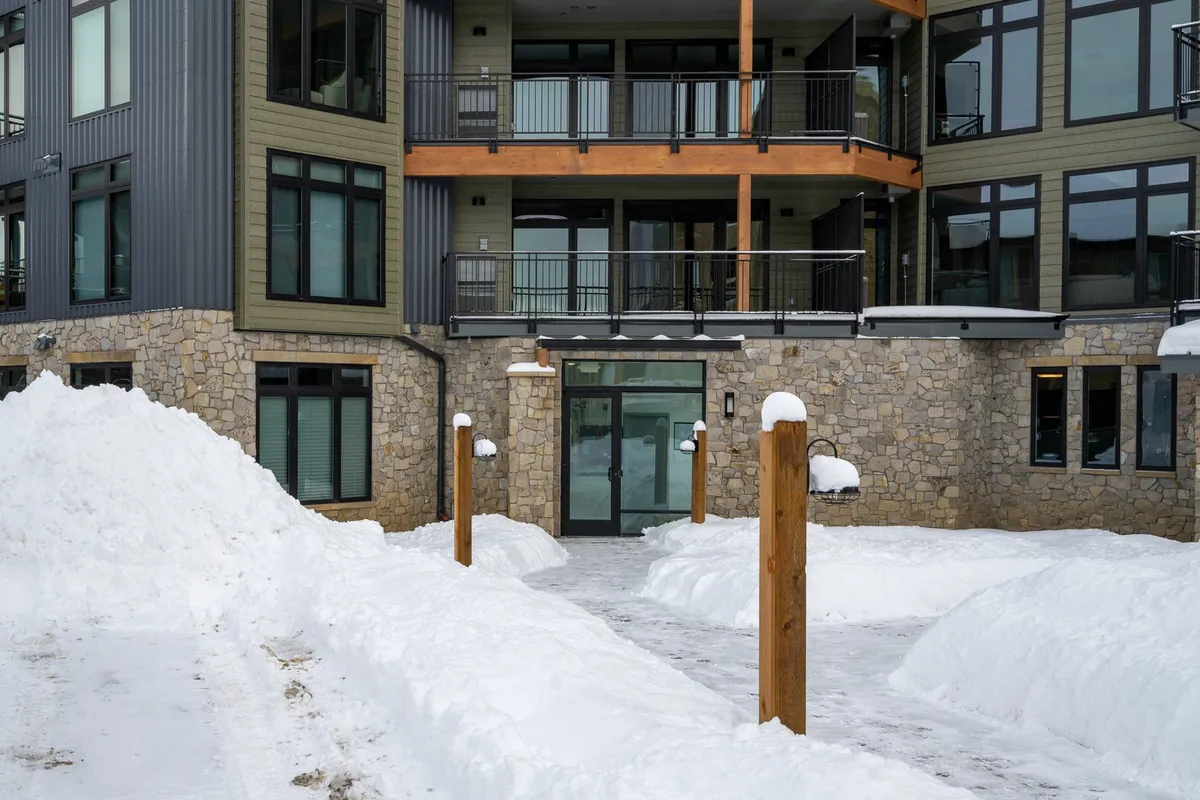 A welcoming exterior scene showing snow-covered pathways, illuminated wooden posts, and the warm stone-and-wood facade of the building. The balconies suggest opportunities for social gatherings and relaxation after days spent exploring.