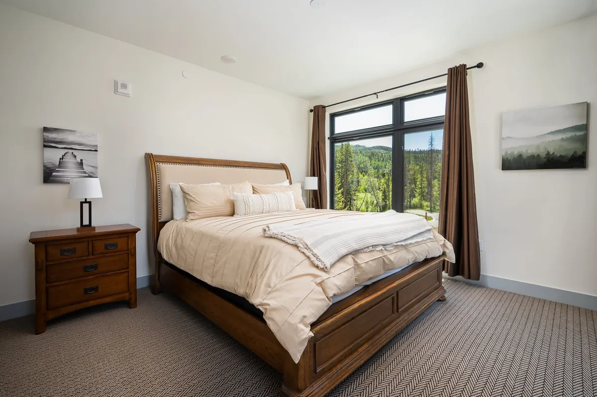 A serene bedroom with a plush bed, large windows letting in ample natural light, a nightstand, and scenic landscape visible outside. The inviting setting is styled for comfort, with soft linens and a calm color palette, ideal for restful nights.