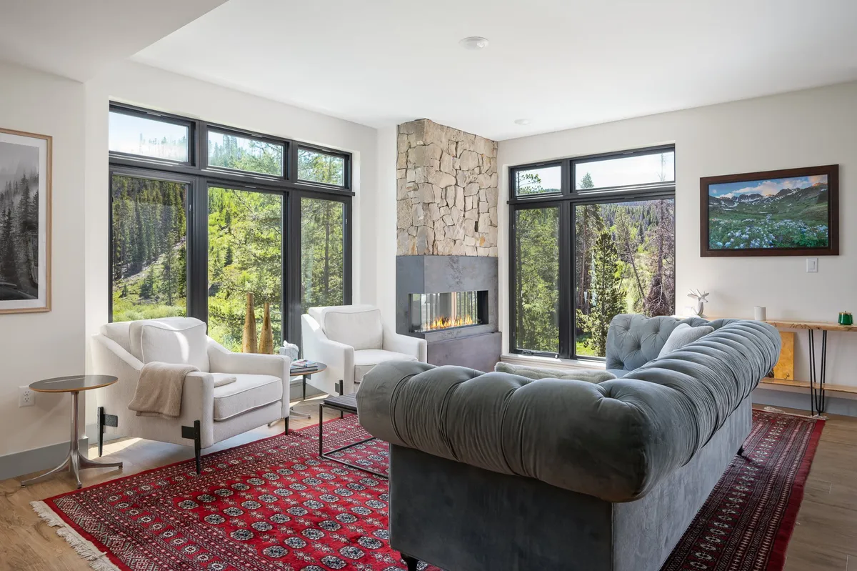 A spacious, inviting living room featuring a plush grey sofa, a striking stone fireplace, and expansive windows framing serene treetop views. Warm natural light floods the space, highlighting cozy seating and a vibrant red-patterned rug, creating a perfect spot for gathering and relaxing after mountain adventures.