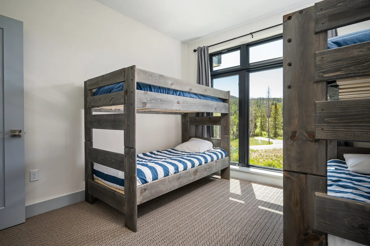 A cheerful bunk room with sturdy bunk beds and a large window letting in natural light. The inviting space is designed for children to sleep, play, or read, with a backdrop of scenic outdoor views.