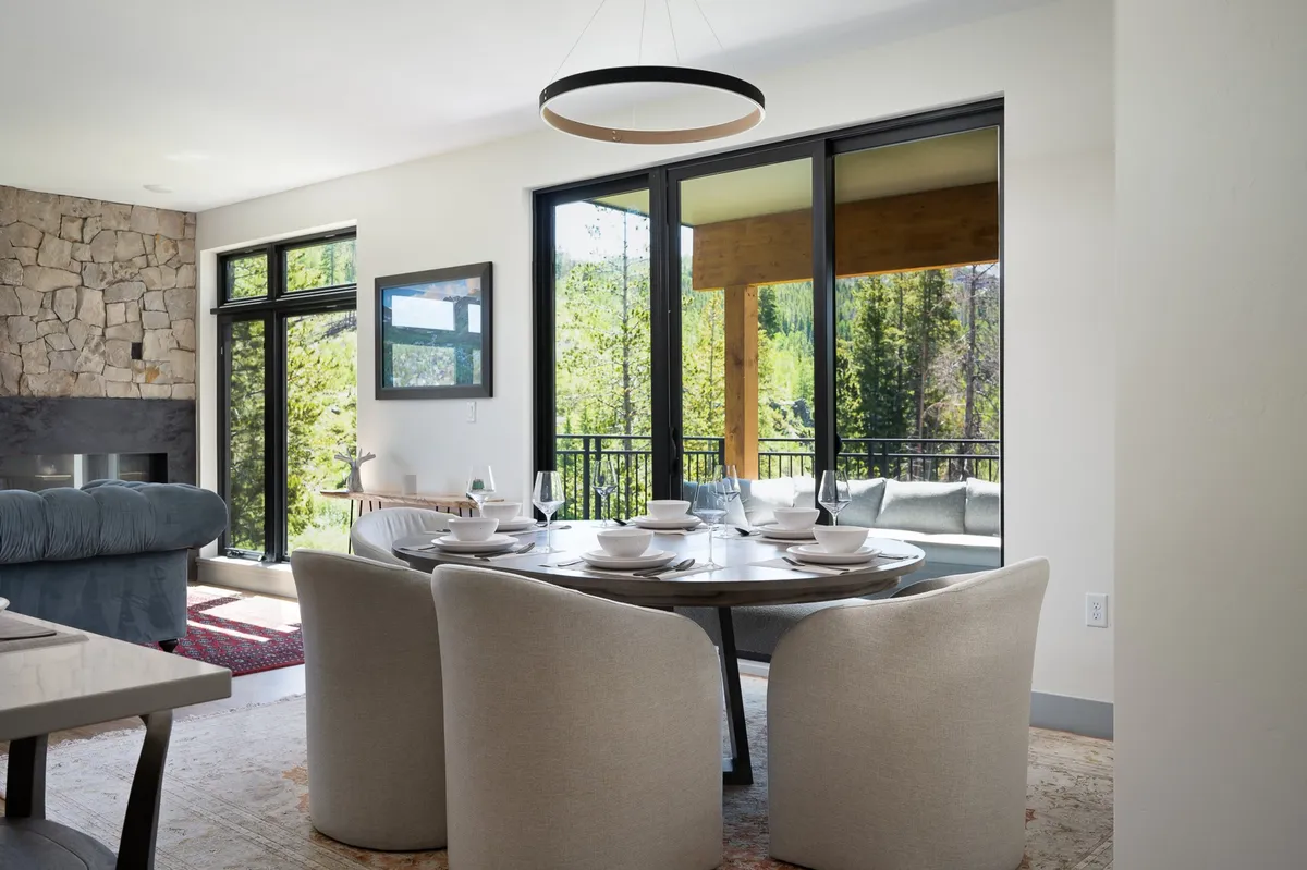 An inviting dining area with a large table set for a meal, flanked by expansive windows that frame serene forest views. The space features a textured stone fireplace, creating a warm atmosphere ideal for both lively dinners and peaceful mornings.