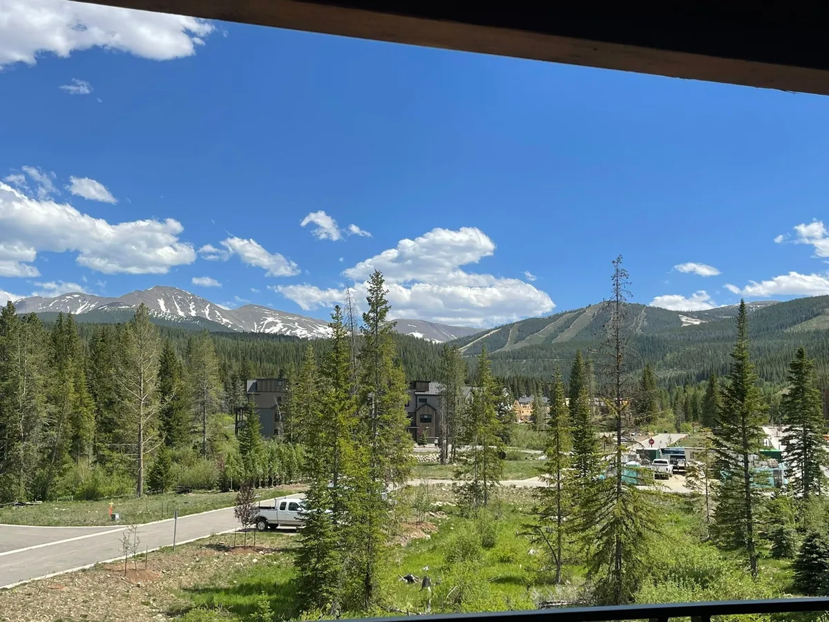 A panoramic view of snow-capped mountains, dense evergreen trees, and newly constructed buildings nestled among the landscape. The clear blue sky and natural grandeur convey a sense of escape and exclusivity.