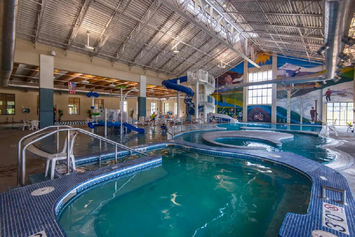 A vibrant view of the Grand Park Rec Center's indoor pool area, featuring water slides, a large swimming pool, mural walls, and seating for family gatherings. The mood is energetic, highlighting fun and connection.