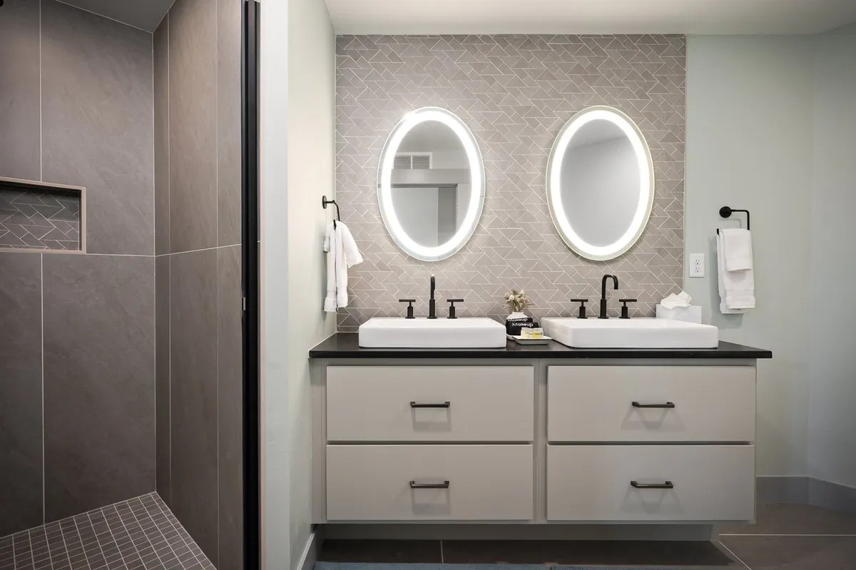 A spa-inspired bathroom with double sinks, elegant mirrored lighting, and a modern shower area. The space exudes a serene, contemporary feel, perfect for relaxation and self-care routines.