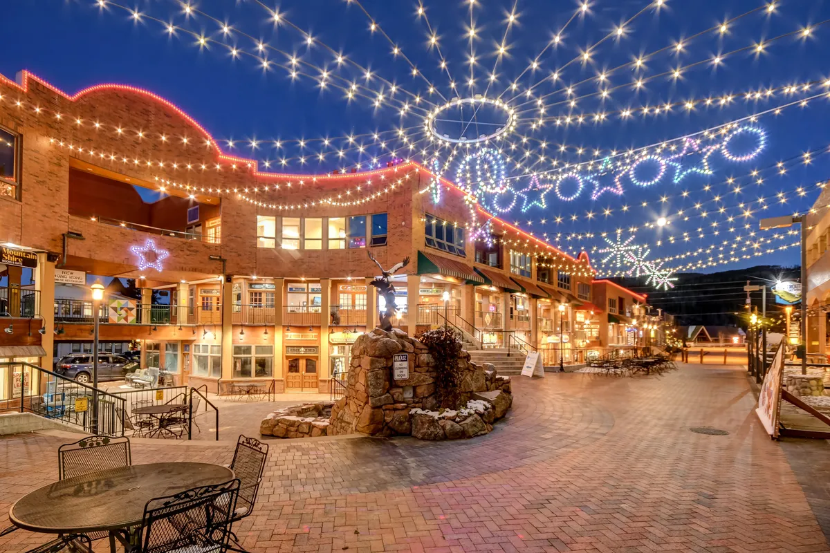 A street-level view of a charming plaza bathed in string lights, with outdoor seating, a decorative fountain, and brick walkways. People casually stroll, dine, and enjoy a festive, communal atmosphere reminiscent of holiday celebrations or evening gatherings.