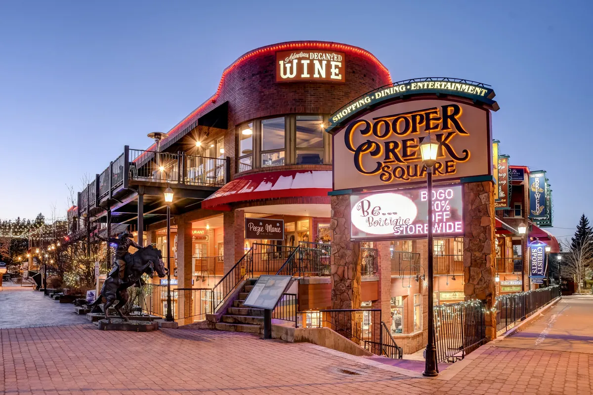 This image captures the vibrant Cooper Creek Square in Downtown Winter Park, featuring festive lighting, a wine store sign, and outdoor seating areas filled with people enjoying the lively atmosphere. Decorative lights and signage hint at thriving local businesses and regular community events.